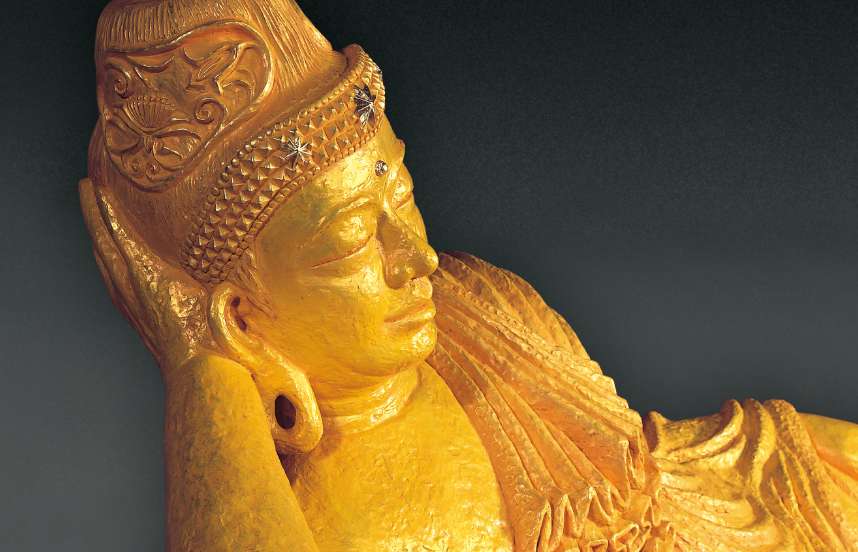 A closeup of the face of a golden colored reclining Buddha sculpture shows a slight smile on his lips; decorative details of the headpiece he wears are visible.