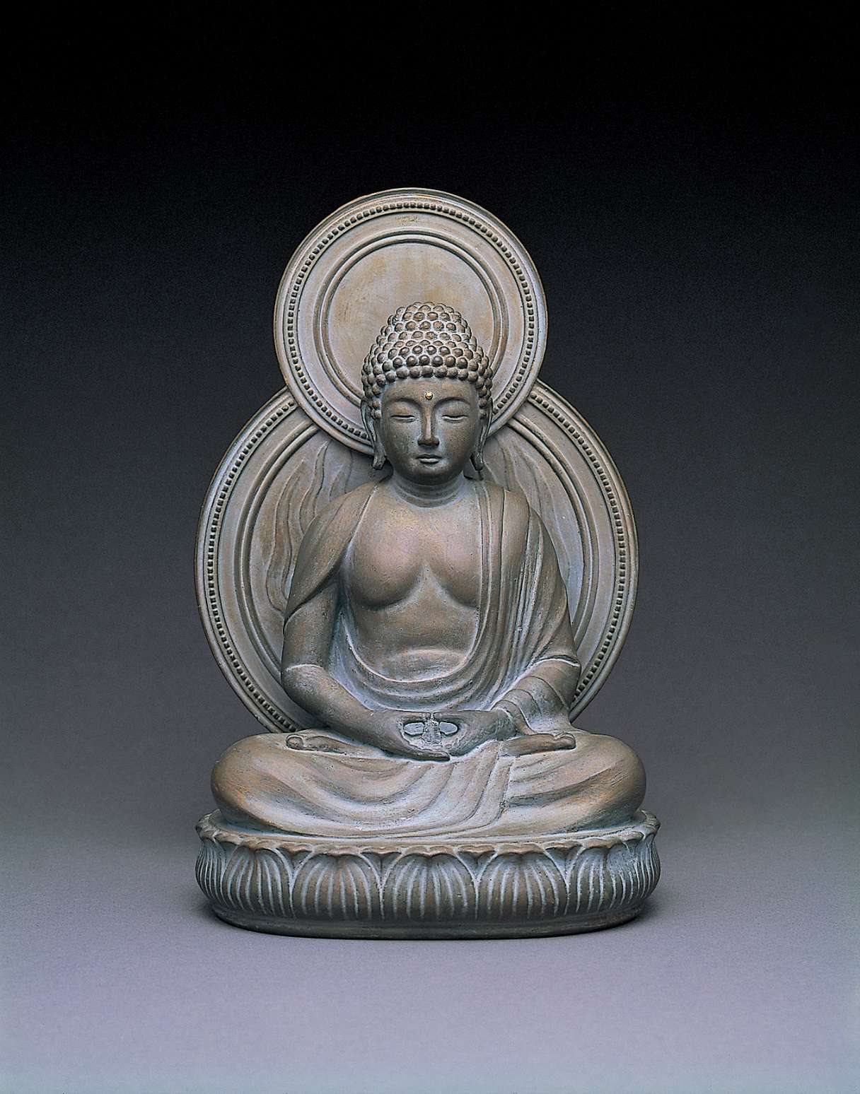 A matte, gray hued statue of a buddha sitting cross-legged atop a seat of curving lotus petals, with a circular nimbus behind his body, hands in meditation.